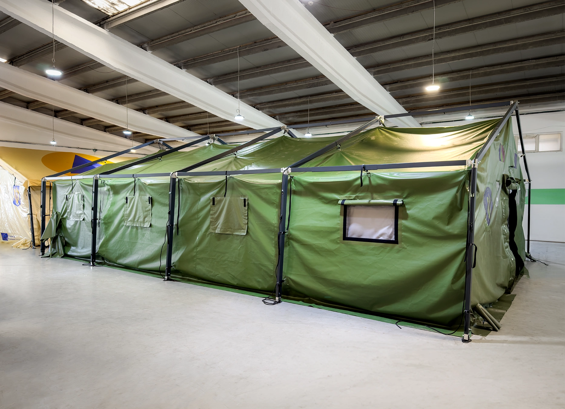 Cort modular militar verde, cu structur? metalic? exterioar? ?i ferestre transparente, montat într-o hal? de produc?ie.