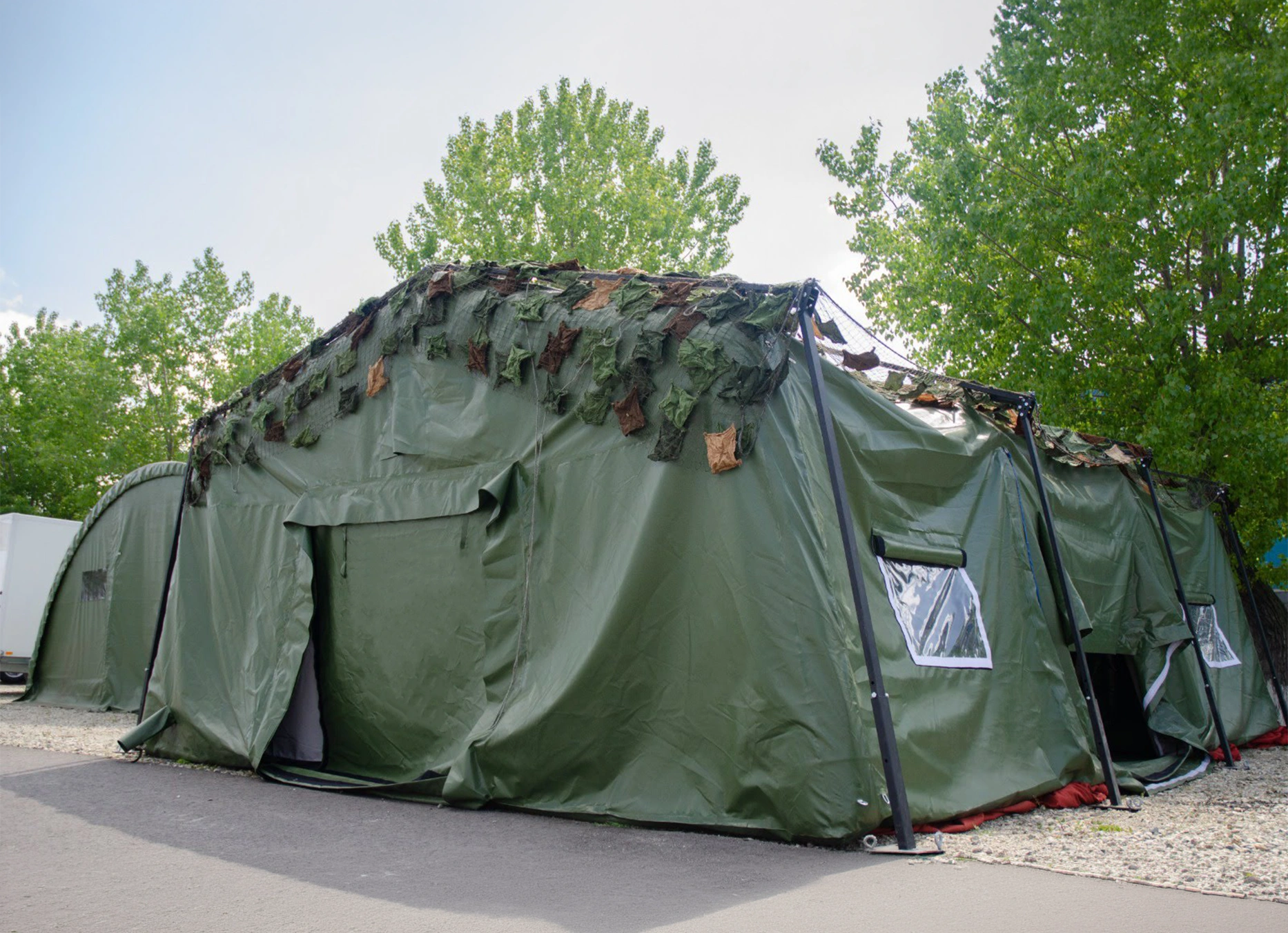 Cort militar modular verde cu structur? metalic?, ferestre transparente ?i plas? de camuflaj, instalat în exterior.